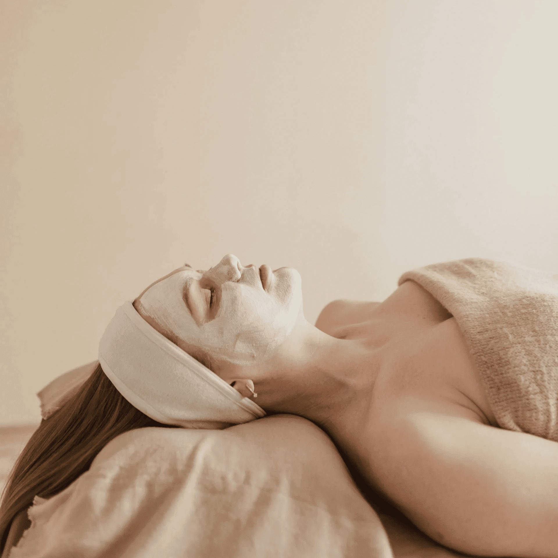 Person relaxing with a white face mask in a spa setting, wrapped in a beige towel.